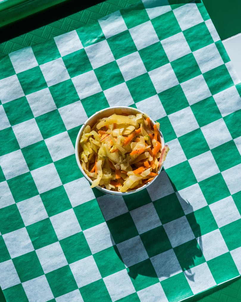 A white paper cup filled with coleslaw, featuring shredded cabbage and carrots, on a green and white checkered paper.
