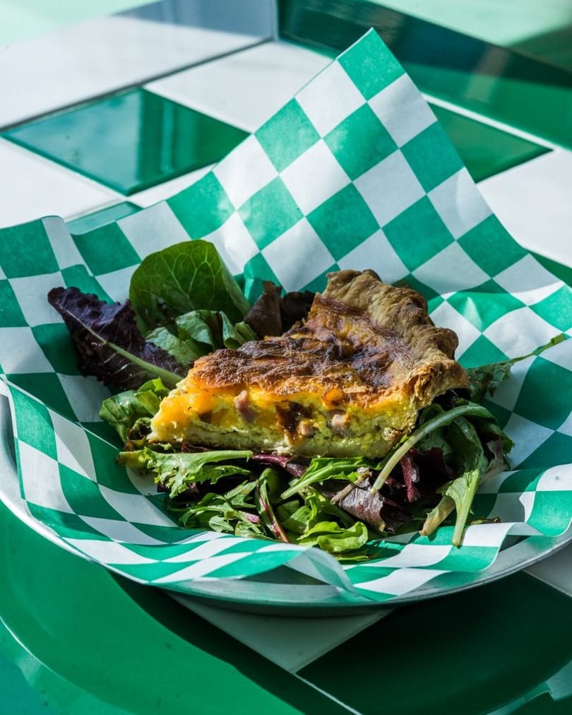 A slice of quiche with a side salad in a green and white checkered paper-lined metal dish.
